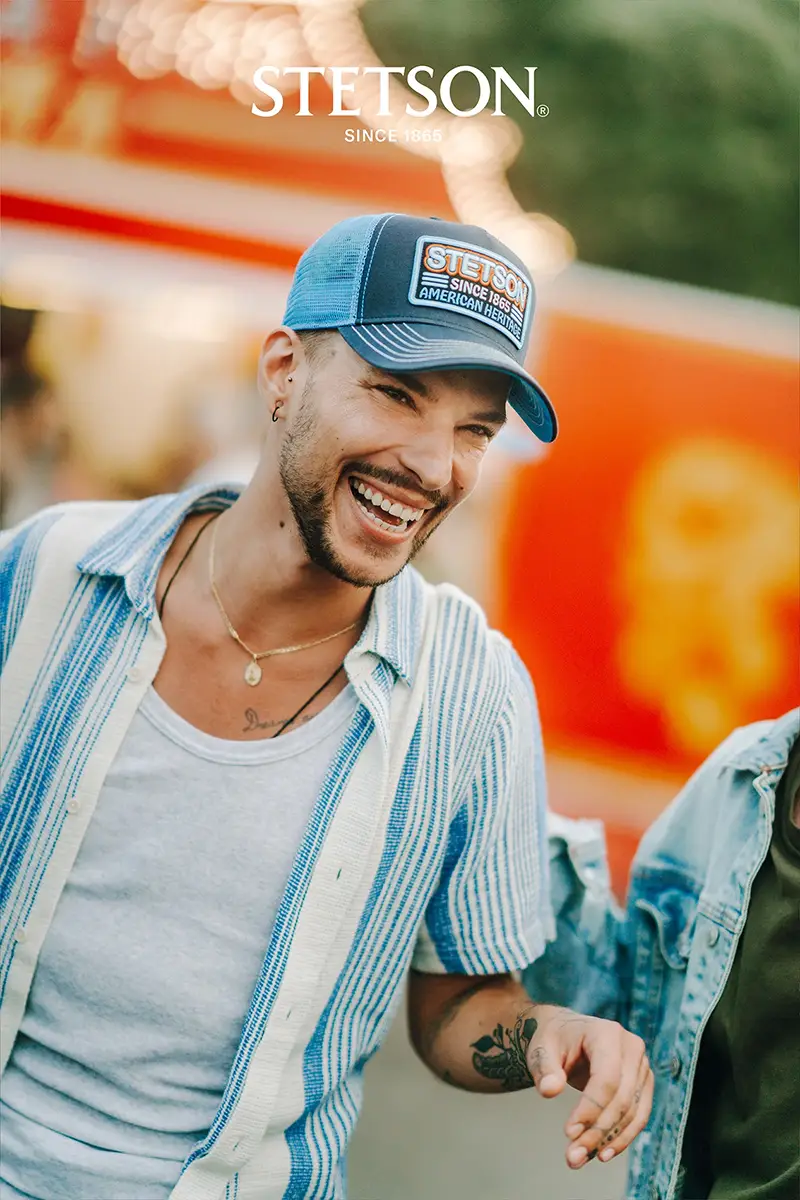 Stetson Trucker Caps – Lässiger Retro-Style mit Markenstatement für den Sommer Fröhlicher junger Mann mit Stetson Trucker Cap im Retro-Stil, lässig gekleidet auf einem bunten Jahrmarkt.